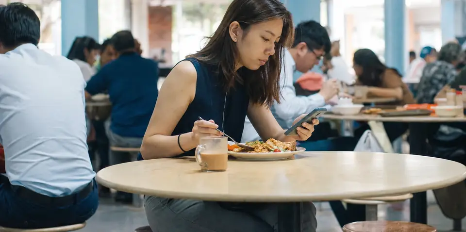 A young woman in office wear having lunch at a hawker centre table, glancing at her phone