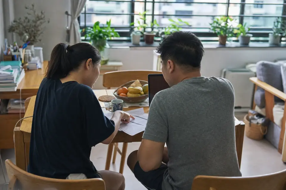 A couple at the dining table of their flat, looking through papers together, with mugs and a fruit bowl