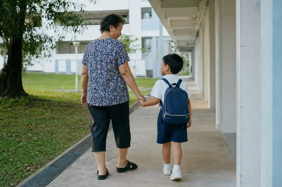 A grandmother walking hand in hand with her young grandson along a sheltered walkway beside HDB blocks